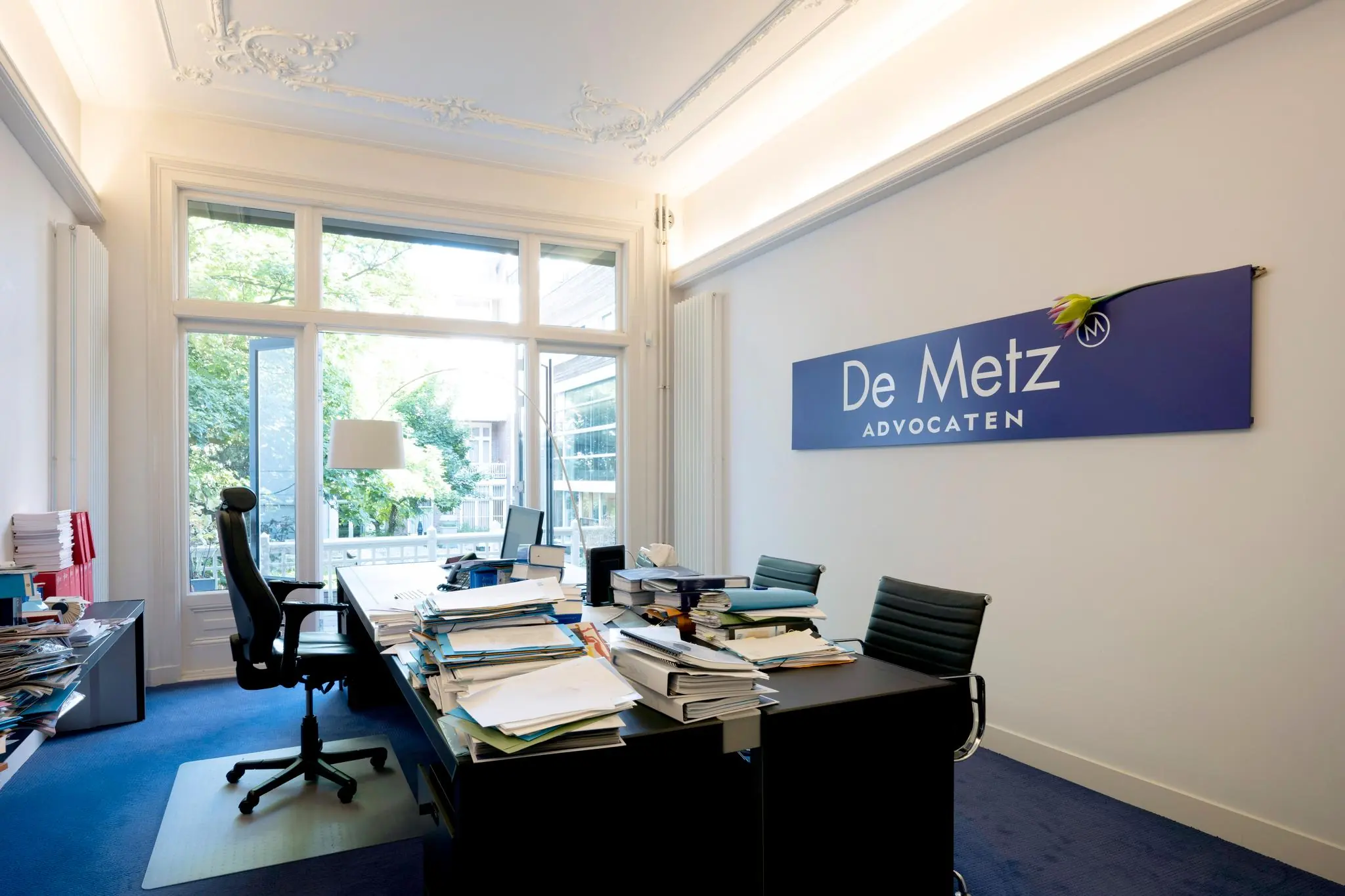 Modern kantoorinterieur van advocatenkantoor De Metz aan de Paulus Potterstraat met een bureau vol dossiers en een groot raam met uitzicht op groen.